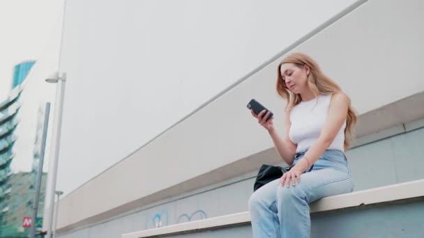 スマートフォンを使って屋外に座っている若い白人女性オンラインショッピングと取引 広告コピースペース — ストック動画