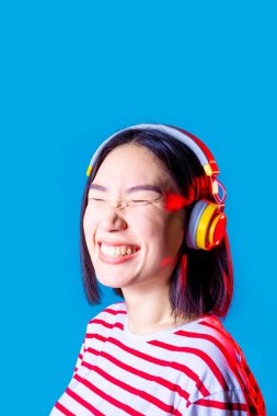 Huzurlu Asyalı kadın mavi arka planda izole edilmiş Bluetooth kulaklıklı, gülümseyen kulaklıklı müzik dinliyor.