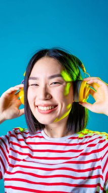 Huzurlu Asyalı kadın mavi arka planda izole edilmiş Bluetooth kulaklıklı, gülümseyen kulaklıklı müzik dinliyor.