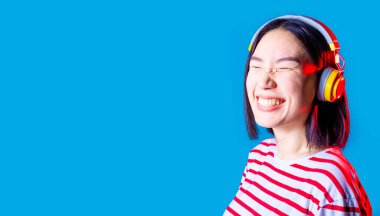 Huzurlu Asyalı kadın mavi arka planda izole edilmiş. Kablosuz kulaklıklar dinliyor. Gülümsüyor.