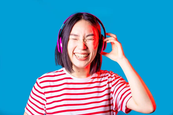 Huzurlu Asyalı kadın mavi arka planda izole edilmiş. Kablosuz kulaklıklar dinliyor. 