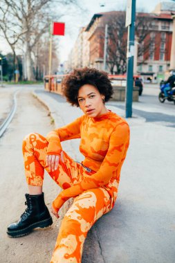 Şehrin dışında poz veren genç melez kadın kameraya ciddi ve kendinden emin görünüyor.