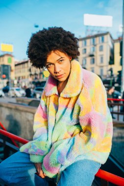 Şehrin dışında poz veren genç melez kadın ciddi ve kendinden emin görünüyor.