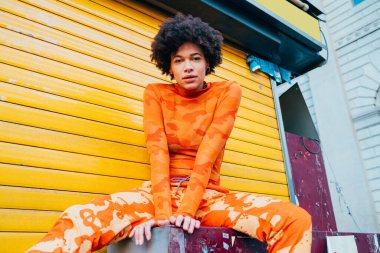 Açık havada poz veren genç melez kadın kameraya ciddi ve kendinden emin bakıyor.