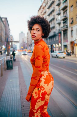 Şehrin dışında poz veren genç melez kadın kameraya ciddi ve kendinden emin görünüyor.