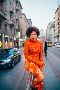 Şehrin dışında poz veren genç melez kadın kameraya ciddi ve kendinden emin görünüyor.