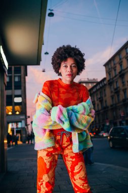 Şehrin dışında poz veren genç melez kadın kameraya ciddi ve kendinden emin görünüyor.