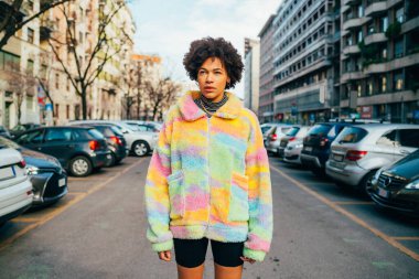 Kendine güvenen, şık, genç siyahi bir kadın. Açık havada havalı bir şekilde poz veriyor. Gülümsüyor, hayal kuruyor ve derin derin düşünüyor.