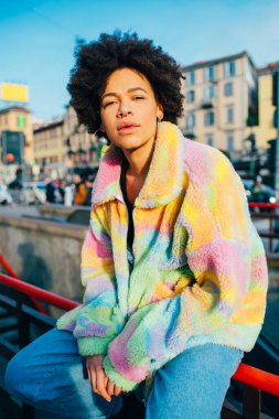 Genç siyah kıvırcık saçlı, şık, havalı, dışarıda poz veren, kameralı, pozitif tavırlı bir kadın.