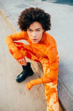 Tarz sahibi genç siyah kadın, kıvırcık saçlı, dışarıda poz veren ciddi ve havalı, kendine güvenen bir kadın.