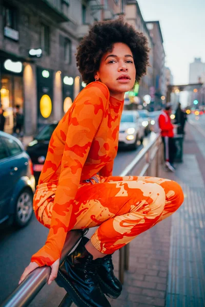 Tarz sahibi genç siyah kadın, kıvırcık saçlı, dışarıda poz veren ciddi ve havalı, kendine güvenen bir kadın.