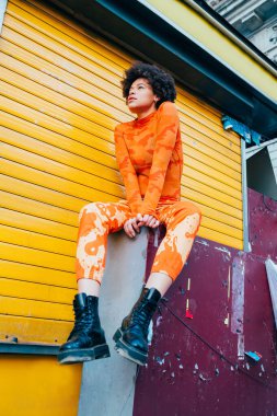 Güçlü ve güçlü genç, siyah stil sahibi, kıvırcık saçlı, açık havada oturmuş, dalgın dalgın bakıyor.