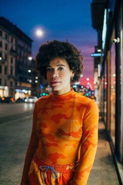 Genç siyah kadın kıvırcık saçlı, şık, şık, açık havada poz veren, kamera görünümlü, havalı tavırlı.