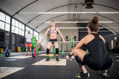 Coach watching young woman athlete deadlift weight lifting barbell training indoors gym