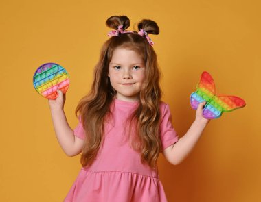 Pembe elbiseli, gülümseyen kızıl saçlı kız elinde iki yeni duyusal gökkuşağı renginde oyuncak tutuyor.