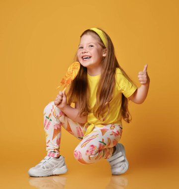 Sarı tişörtlü ve renkli taytlı gülen kızıl saçlı kız çömelmiş, lolipop tutuyor, baş parmak işareti yapıyor.