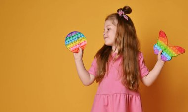 Pembe elbiseli kızıl saçlı kız elinde iki duyusal gökkuşağı rengi ve kelebek şeklinde oyuncaklar tutuyor.