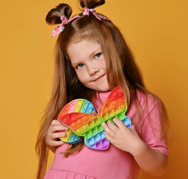Red-haired kid girl in pink shirt stands hugging two new sensory rainbow color toys - butterfly shape and round pop it — Stock Photo, Image