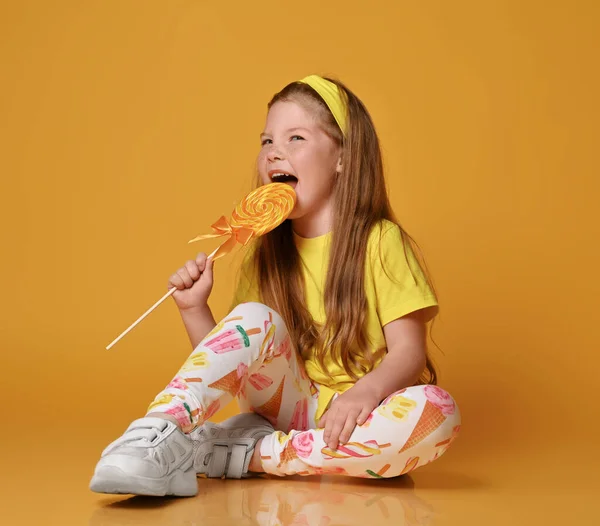 Frolic niña de pelo rojo en camiseta amarilla y polainas de colores se sienta en el suelo, comiendo piruleta y mira hacia arriba de ensueño Imágenes de stock libres de derechos