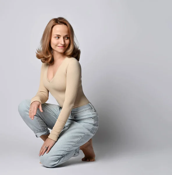 Young blonde woman with in beige bodysuit with long sleeves, shirt and blue jeans sits squatting and looks behind back — Stock Photo, Image