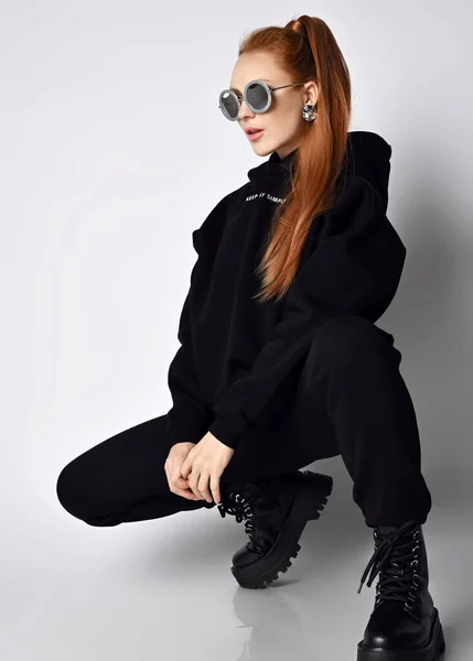 Smala fashionabla rödhåriga kvinna i trendiga svarta sportkläder hoodie, byxor och massiva brutala skor sitter knäböjda i sidled — Stockfoto