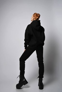 Süslü kapüşonlu, pantolonlu ve vahşi ayakkabılı genç seksi kadın kameraya arkası dönük duruyor.