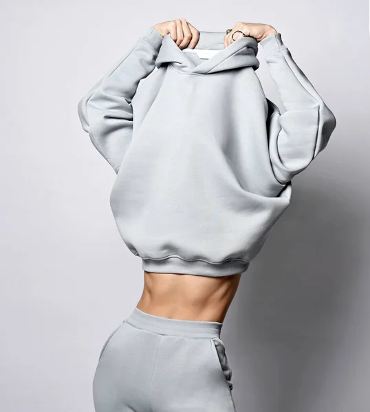 Slim fitness woman in grey sportswear takes off her hoodie, undresses, demonstrating her waistline and effect of diet — Stock Photo, Image