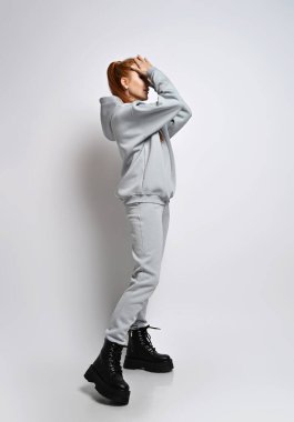 Sıska, kızıl saçlı, moda gri kapüşonlu, pantalonlu, siyah ayakkabılı kadın alnının önünde el ele duruyor.