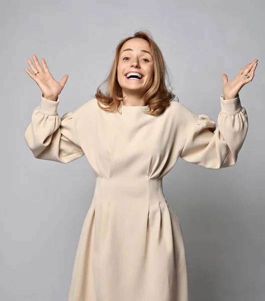 Portrait of happy smiling, ecstatic emotional young blonde woman in beige dress stands holding hands up and laughs — Stock Photo, Image