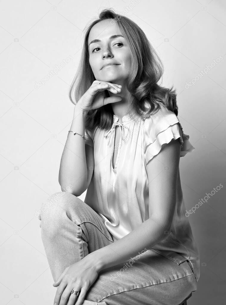 Black and white portrait of young woman with long blonde hair in blouse and jeans sitting squatted looking up dreamily
