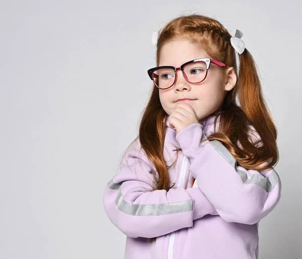 Retrato de menina criança ruiva inteligente em óculos e macacão rosa com capuz de pé segurando a mão em seu queixo, pensando — Fotografia de Stock