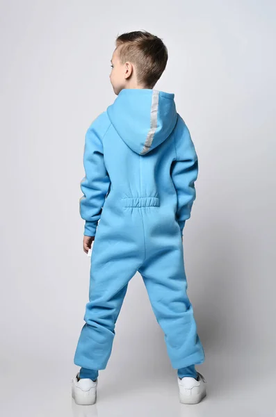 Mały chłopiec w niebieskim kombinezonie z kapturem z paskiem odblaskowym stoi tyłem do kamery z odwróconą głową i patrząc w bok — Zdjęcie stockowe