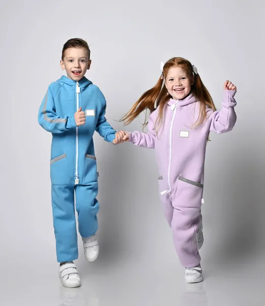 Zwei Jungen und Mädchen in blauen und rosa Jumpsuits mit Reißverschlüssen und Taschen laufen händchenhaltend auf die Kamera zu lizenzfreie Stockfotos