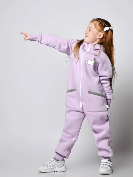 Enfant fille Frolic en combinaison moderne rose se tient sur le côté pointant avec le doigt au coin supérieur, de côté — Photo