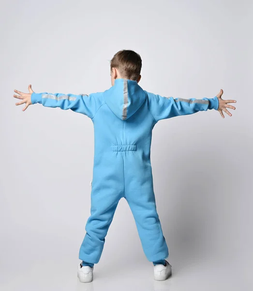 Kid boy in blauw jumpsuit met capuchon met reflecterende streep staat terug naar camera met armen wijd uit elkaar. Achteraanzicht — Stockfoto