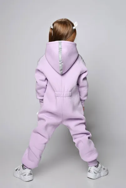 Portrait of redhead kid girl in pink jumpsuit with hood standing back to camera with legs wide apart, hands in pockets — Stock Photo, Image