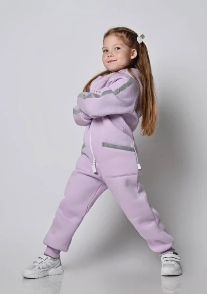 Frolic proudful kid girl in pink modern jumpsuit stands with legs wide apart, arms crossed at chest Stock Image