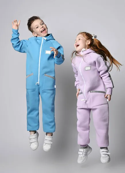 Lekfulla barn pojke och flicka i blå och rosa overall hoppar tillsammans, har kul, skrattar högt Royaltyfria Stockbilder