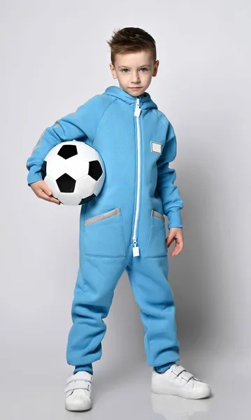 Веселый, активный мальчик в синем комбинезоне с капюшоном и карманами с отражающими полосками стоит, держа футбольный мяч — стоковое фото