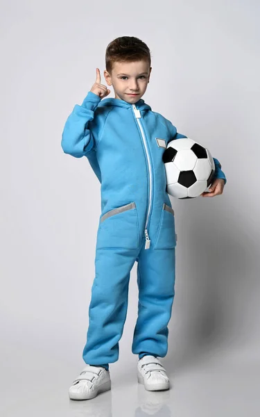 Garoto de brincadeira em macacão azul com bolsos fica segurando bola de futebol na mão e gesticulando dedo indicador para cima — Fotografia de Stock