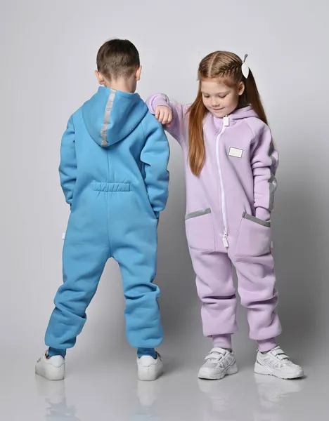 Two kids in jumpsuits are standing next to each other, boy with his back to us and girl with her face and looks down Stock Photo