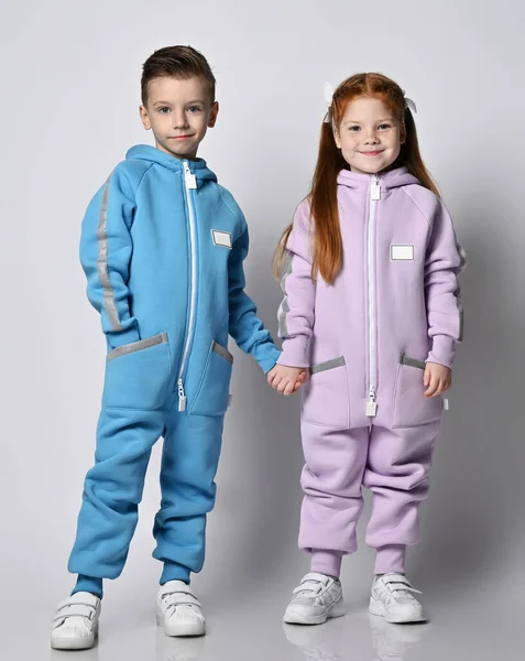 Tow cute smiling kids boy and girl in blue and pink jumpsuits with hoods and pockets stand together holding hands — Stock Photo, Image
