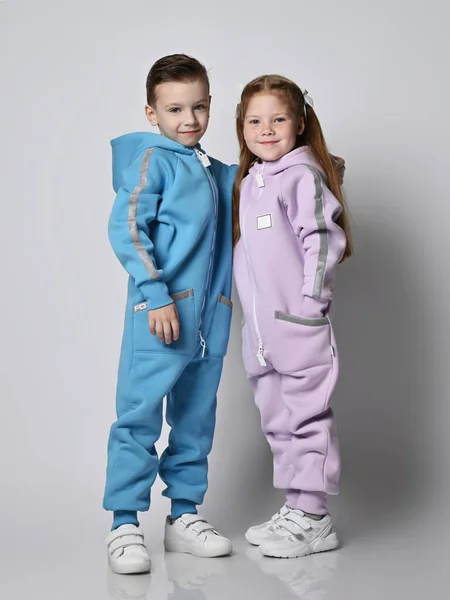 Im Schlepptau lächelnde Kinder Junge und Mädchen, Freunde in blauen und rosa Jumpsuits mit Kapuzen und Taschen stehen dicht beieinander. Stockfoto