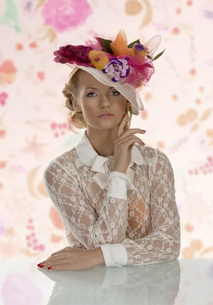 Menina elegante atrás da mesa com chapéu floral e mão perto do rosto — Fotografia de Stock