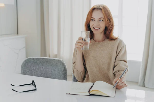 Glad rödhårig kvinna med glas rent mineralvatten skriva inköpslista i bärbar dator hemma — Stockfoto