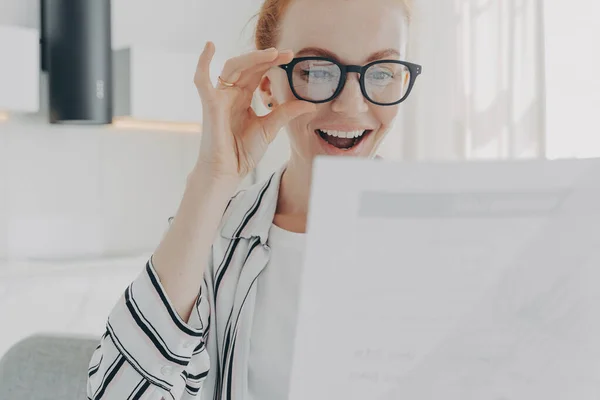 Ruiva positiva jovem mulher sorri amplamente usa óculos transparentes focados em papéis — Fotografia de Stock