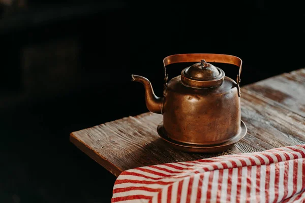 Горизонтальний знімок античного чайника на старовинному дерев'яному столі. Старий посуд. Металевий мідний чайник на фоні темного червоного рушника поруч. Вибірковий фокус — стокове фото