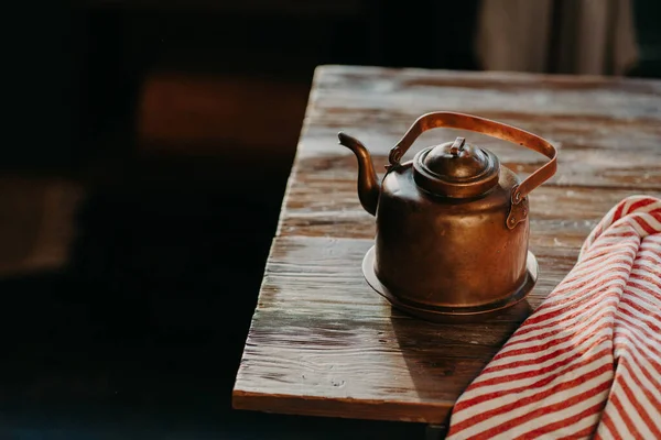 Старий мідний металевий чайник на дерев'яному столі в темній кімнаті. Поруч червоний смугастий рушник. Античний чайник для приготування чаю або кави. Кухонне обладнання — стокове фото