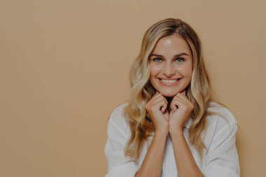 Genç sarışın kadın ellerini çenesinin altında kavuşturuyor ve kameraya sinsice bakıyor.