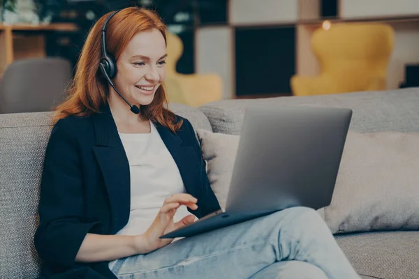 Ung positiv gingerhead kvindelig studerende har online webinar via laptop og headset i kaffebar - Stock-foto